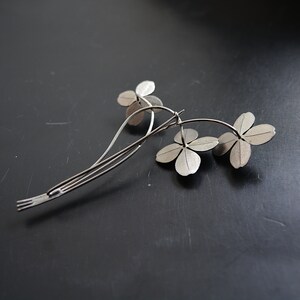 May include: A silver-toned hair clip featuring a floral design with three four-leaf clover blooms. The clip has a slender, curved stem and a barrette-style clasp. The metal has a matte finish, and the piece is set against a dark background.