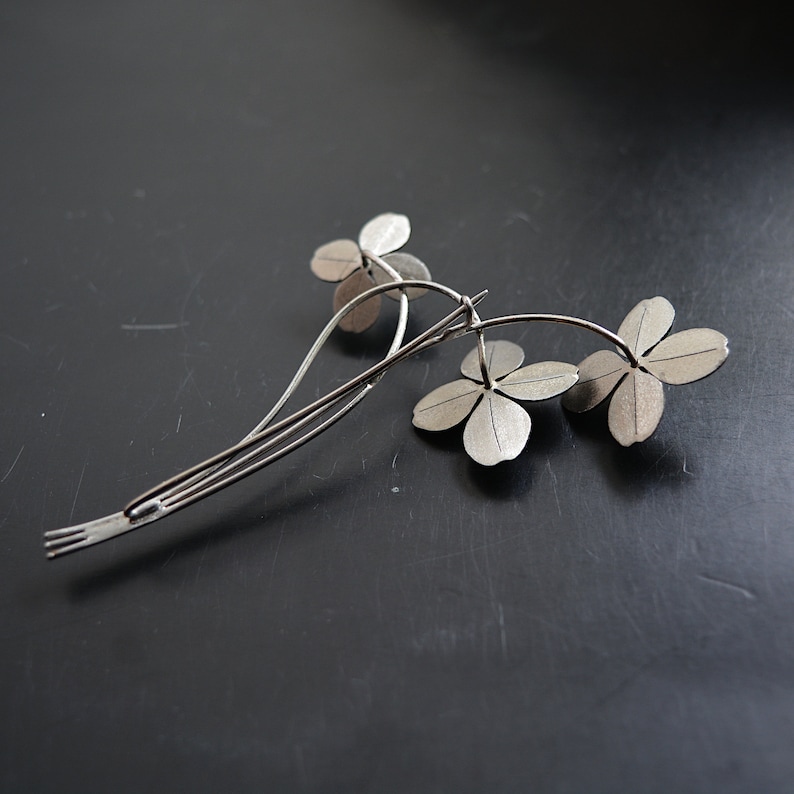 May include: A silver-toned hair clip featuring a floral design with three four-leaf clover blooms. The clip has a slender, curved stem and a barrette-style clasp. The metal has a matte finish, and the piece is set against a dark background.