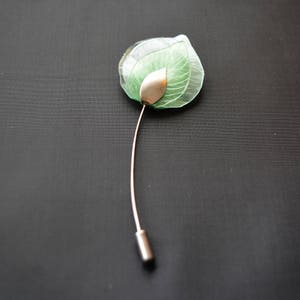 May include: A green glass flower brooch with a silver leaf detail. The brooch is attached to a silver pin.