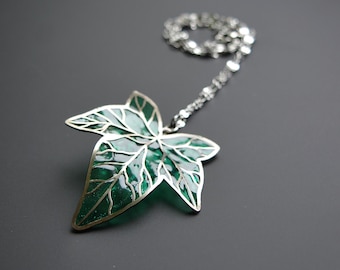 May include: A silver-toned necklace featuring a green leaf pendant. The leaf design has a detailed, veined pattern with a glossy, emerald green fill. The chain is delicate and attached to the top of the leaf.