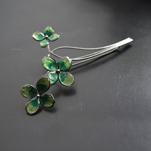 May include: A silver-toned hair accessory featuring a floral design with four-leaf clover-shaped elements. The clover leaves are a translucent green, and the accessory has a delicate, elegant appearance.