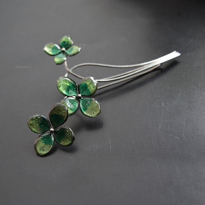 May include: A silver-toned hair accessory featuring a floral design with four-leaf clover-shaped elements. The clover leaves are a translucent green, and the accessory has a delicate, elegant appearance.