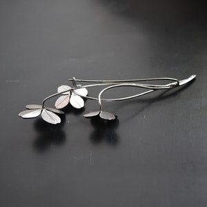 May include: A silver clover brooch featuring four leaves and a delicate stem. The brooch is crafted from metal and has a simple, elegant design.