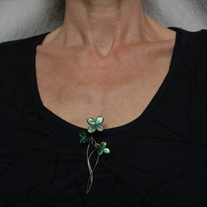 May include: A silver brooch featuring three green flower-shaped charms. The flowers have a metallic sheen and are arranged in a cluster.