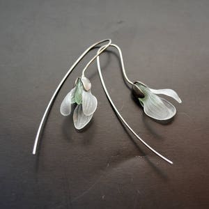 May include: A pair of silver earrings with delicate white flower charms. The flowers have a translucent, almost paper-like texture and are accented with green details.