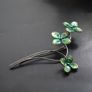 May include: A silver-toned brooch featuring a floral design with three clusters of green, four-leaf clover-like shapes. The leaves have a mottled, glossy finish, and the brooch has a delicate, wire-like stem.
