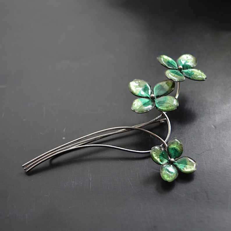 May include: A silver-toned brooch featuring a floral design with three clusters of green, four-leaf clover-like shapes. The leaves have a mottled, glossy finish, and the brooch has a delicate, wire-like stem.