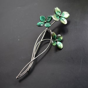 May include: A silver brooch featuring three green enamel flowers. The flowers are made of four petals each and have a slightly textured surface.