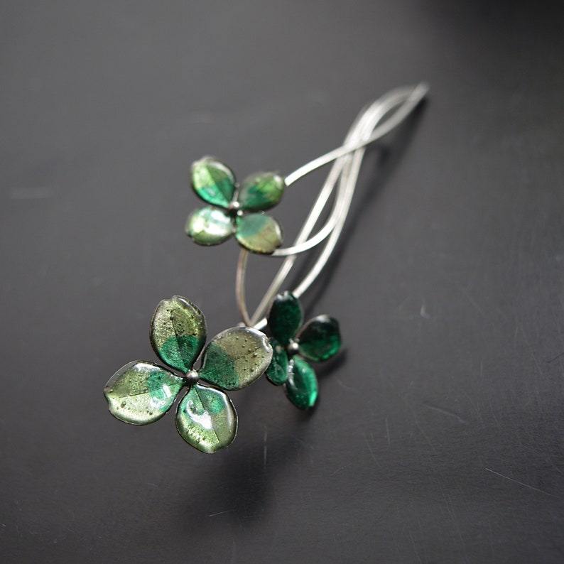 May include: A silver hair comb with three green glass flowers. The flowers have a layered look with a darker green centre and a lighter green outer layer.