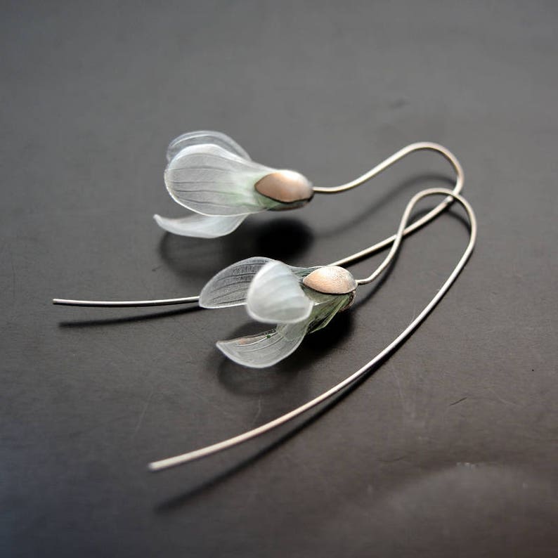 May include: A pair of delicate silver earrings with white flower-shaped charms and a subtle green tint. The earrings have a simple, elegant design.