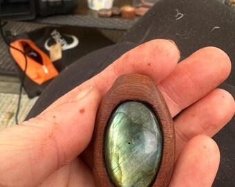 May include: A wooden pendant with a green and blue labradorite gemstone set in the center. The pendant is oval shaped and has a dark brown wood frame.