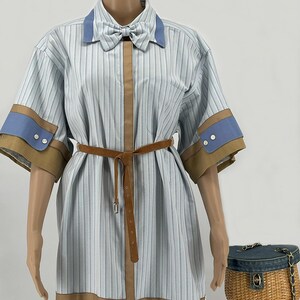 May include: A light blue and white striped dress with a brown belt and bow tie. The dress has short sleeves with blue and brown accents. A small denim purse with a wicker base sits to the side.
