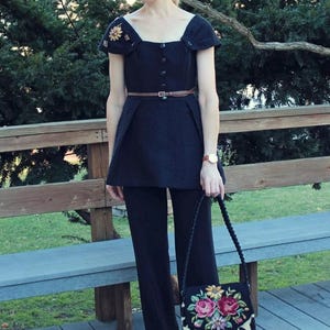 May include: A woman's black outfit, including a top with floral embellishments, a brown belt, and matching pants. She carries a black purse with a floral design and a leopard print flap. The outfit is accessorized with sunglasses and a watch.