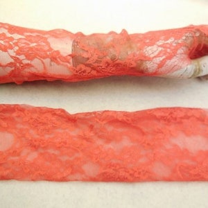 May include: A pair of coral lace fingerless gloves. The gloves are made of a delicate, sheer lace fabric with a floral pattern. The gloves are shown laid flat and on a hand.