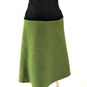 May include: A knee-length skirt featuring a solid black high-waisted band and a textured olive green lower section. The garment has a simple A-line silhouette with a clean, straight hemline, showcasing a clear contrast between the dark waistband and the vibrant green fabric body.