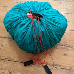 Könnte beinhalten: Ein türkisfarbener Fallschirmbeutel mit roten Kordeln und schwarzen Riemen. Der Beutel ist mit einem Fallschirm gefüllt.