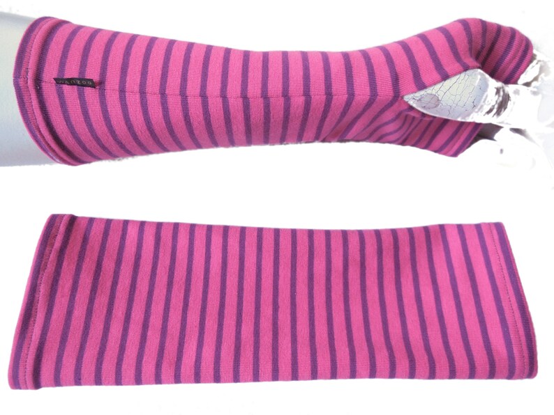 May include: Pink and purple striped arm warmers. The arm warmers are made of a soft, stretchy fabric and feature a repeating pattern of vertical stripes. The arm warmers are shown in two views, one on a mannequin arm and one flat.