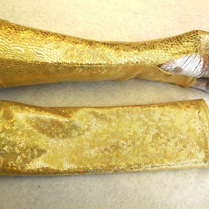 Puede incluir: Un par de guantes largos sin dedos de color dorado con una textura escamosa. Los guantes están hechos de un material brillante e iridiscente.