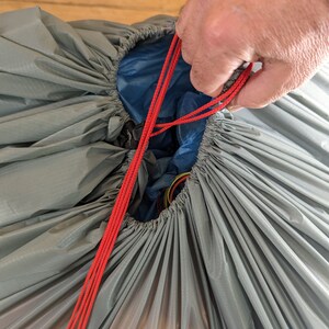 Op de afbeelding: Close-up van een grijze nylon tas met een trekkoordsluiting. Felrode koorden worden strakgetrokken en trekken de opening samen. De binnenkant onthult een blauwe voering en kleurrijke draden.