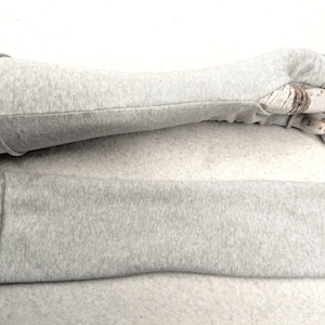 Puede incluir: Un guante gris claro sin dedos con una manga a juego, exhibido en una mano de maniquí. El guante está hecho de una tela de punto suave. También se muestra una pieza rectangular separada de la misma tela.