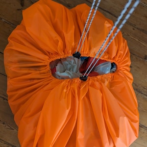 Op de afbeelding: Fel oranje stoffen tas, aan de bovenkant gerimpeld met witte en zwarte koorden. De tas is gedeeltelijk open en toont de inhoud. Hij ligt op een houten oppervlak.