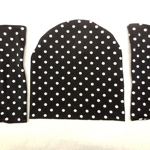 May include: Three pieces of black fabric with white polka dots. The pieces include a beanie-style hat and two matching arm warmers. The fabric is a soft material, perfect for a cozy accessory set.