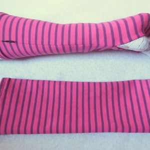 May include: Pink and purple striped arm warmers. One arm warmer is on a mannequin arm, the other is lying flat. The arm warmers are made of a soft fabric and have a thumb hole. The stripes are vertical and evenly spaced.