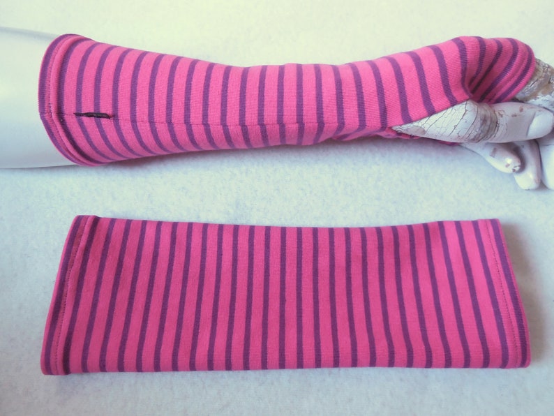 May include: Pink and purple striped arm warmers. One arm warmer is on a mannequin arm, the other is lying flat. The arm warmers are made of a soft fabric and have a thumb hole. The stripes are vertical and evenly spaced.
