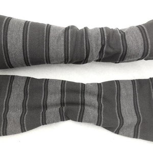 Könnte beinhalten: Ein Paar Armstulpen mit einem sich wiederholenden Muster aus breiten anthrazitfarbenen Streifen, die sich mit dünneren hellgrauen Streifen abwechseln. Der Stoff hat eine weiche Strickstruktur und eine röhrenförmige Form vom Handgelenk bis zum Unterarm.