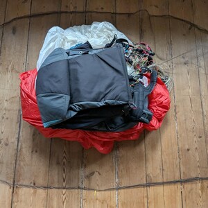 May include: A black and gray paragliding harness and a red and white parachute canopy are laid out on a wooden floor.