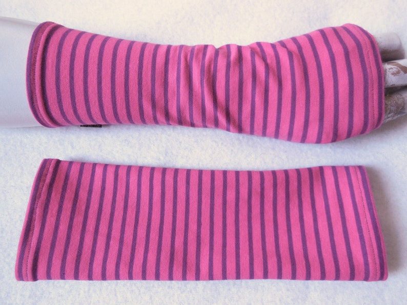 May include: A pair of pink and purple striped arm warmers. One arm warmer is shown on a mannequin arm, while the other is laid flat. The arm warmers are made of a soft, stretchy fabric and feature vertical stripes.