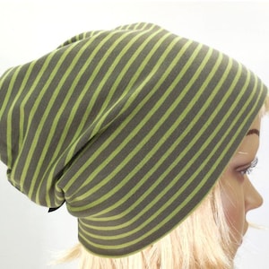 Könnte beinhalten: Eine lockere Beanie-Mütze mit olivgrünen und hellgrünen Streifen. Die Mütze ist aus einem weichen Stoff gefertigt und für den lässigen Gebrauch konzipiert. Die vertikalen Streifen erzeugen ein optisch ansprechendes Muster. Die Mütze wird auf einem Schaufensterkopf gezeigt.