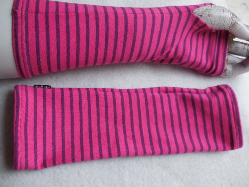 May include: A pair of pink and purple striped fingerless gloves. The gloves are made of a soft material and feature a ribbed cuff. The brand name "WARGO" is on a small black tag. The gloves are designed to cover the hand and forearm.
