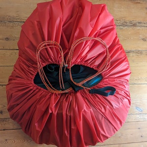 May include: A red, drawstring bag with a black strap and orange and black cords. The bag is lying flat on a wooden surface.