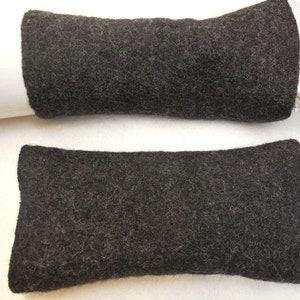 May include: A pair of dark charcoal gray wrist warmers crafted from a thick, felted wool material. These rectangular accessories feature a dense, textured surface and a simple, seamless design that covers the forearm area without any thumb holes.