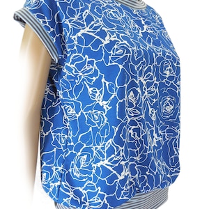 Puede incluir: Una camisa de manga corta con estampado floral azul y blanco con un ribete a rayas alrededor del cuello y las mangas. La camisa tiene un corte holgado y un dobladillo ligeramente fruncido.