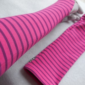 May include: A pair of pink and purple striped arm warmers. The arm warmers are made of a soft, ribbed knit fabric. The stripes are horizontal and evenly spaced. One arm warmer is on a hand, the other is lying flat.