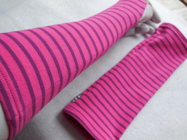 May include: A pair of pink and purple striped arm warmers. The arm warmers are made of a soft, ribbed knit fabric. The stripes are horizontal and evenly spaced. One arm warmer is on a hand, the other is lying flat.