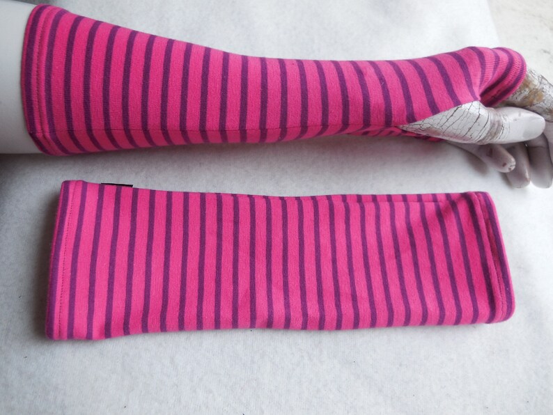 May include: A pair of pink and purple striped arm warmers. One arm warmer is on a mannequin arm, and the other is lying flat. The arm warmers are made of a soft, stretchy fabric and have a thumb hole.
