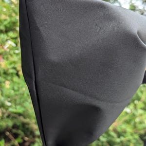 Könnte beinhalten: Eine schwarze, dreieckige Abdeckung, möglicherweise für einen Fahrradspiegel, aus einem strukturierten Stoff. Die Abdeckung ist gefaltet und genäht, mit einer sichtbaren Naht. Der Hintergrund ist unscharf und zeigt grünes Laub.