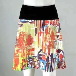 May include: A-line skirt with a black waistband and a colorful abstract print. The print features red, yellow, blue, and white shapes resembling a cityscape. The skirt is knee-length and made of a lightweight fabric.