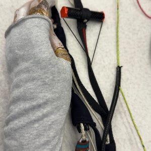 May include: A light gray fabric sleeve with a thumb hole opening, positioned alongside a complex arrangement of black nylon webbing straps, thin white and neon yellow cords, and a small black and orange mechanical toggle component.