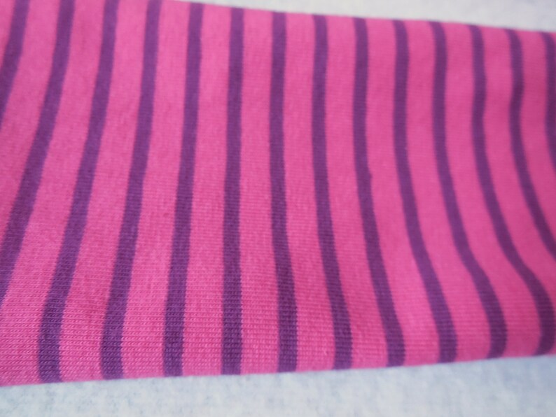 May include: Close-up of a fabric with vertical stripes. The stripes alternate between a bright pink and a deep purple color. The fabric appears to be soft and possibly knitted.