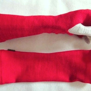 May include: Two bright red arm warmers. One is on a mannequin hand, showing the wrist and thumb opening. The other is a flat, rectangular sleeve. Both are made of a soft, textured fabric. A small tag is visible on the flat sleeve.