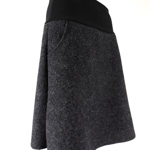 K&ouml;nnte beinhalten: Ein grauer Wollrock mit einem schwarzen elastischen Bund. Der Rock hat eine leicht ausgestellte Form und eine einzelne Tasche auf der Vorderseite.