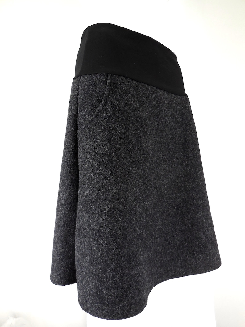 K&ouml;nnte beinhalten: Ein grauer Wollrock mit einem schwarzen elastischen Bund. Der Rock hat eine leicht ausgestellte Form und eine einzelne Tasche auf der Vorderseite.