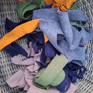 May include: A collection of felt fabric strips in various colours including orange, olive green, navy blue, light blue, and dusty pink. The strips have a textured, matte surface and are arranged in a pile, showing different lengths and widths.