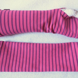 May include: A pair of pink and purple striped arm warmers. The arm warmers are made of a soft fabric and feature vertical stripes. One arm warmer is on a white mannequin arm, and the other is lying flat.