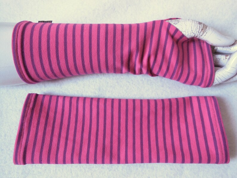 May include: A pair of pink and purple striped arm warmers. The arm warmers are made of a soft fabric and feature vertical stripes. One arm warmer is on a white mannequin arm, and the other is lying flat.