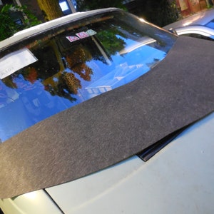 May include: A dark gray, curved car windshield sunshade rests on the hood of a light-colored car. The sunshade is made of a textured material and is designed to fit the curve of the windshield. The windshield reflects the surrounding environment.